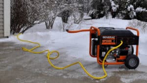 A Generac Portable Generator on a driveway after a winter storm powers essentials during an outage. A heavy duty generator cord runs from the generator to the home's transfer switch inlet box.