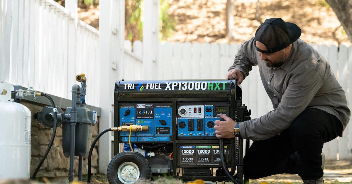 A man starts a DuroMax 13000 Watt Tri Fuel Generator Connected to a propane tank and a manual transfer switch. Backyard Setting with Fence and Deck.
