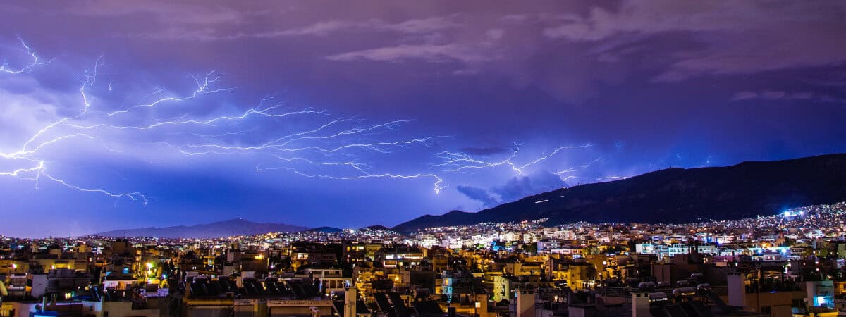 Portions of a large city experience a power outage. Lightning strikes across the sky reach for towers on a hilltop on the right