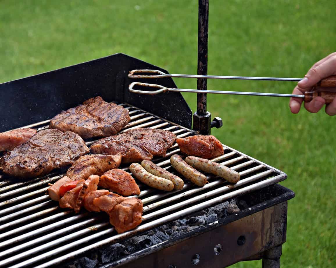 Grilling Steaks, Kabobs, and Sausages on a backyard grill over charcoal. Green grass in the background. A tongs is visible held by a hand