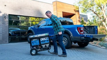 A man wheels a 13000 Watt Generator away from a pickup truck across a paver driveway in front of a modern home with stone and wood siding.